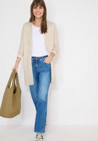 Femme souriante marchant, portant un cardigan beige, une chemise blanche, un jean bleu, des baskets blanches et tenant un grand sac vert olive.
