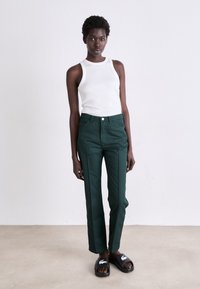 Pantalons en coton verts avec une taille haute et des jambes droites, associés à un haut sans manches blanc et des sandales noires à enfiler.