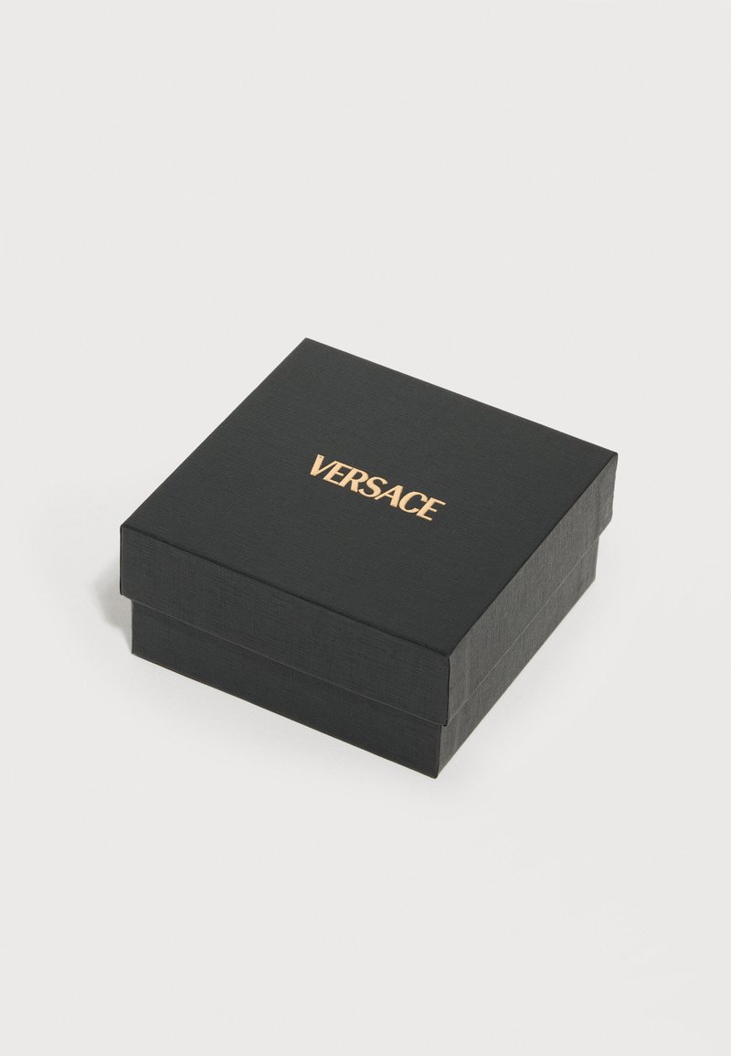 Boîte carrée noire texturée avec le logo VERSACE doré centré sur le couvercle, placée sur une surface blanche.