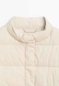 Collo della giacca imbottita beige con scollo tondeggiante. Tessuto liscio e leggermente testurizzato, caratterizzato da un sottile motivo trapuntato e chiusure nascoste.