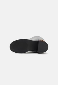 Semelle en caoutchouc noire d'une botte, présentant une surface texturée et une tige grise avec un accent décoratif en chaîne dorée le long du côté.