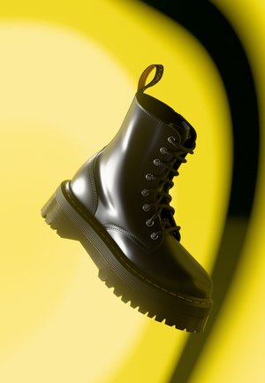 Botte de combat noire brillante à lacets avec semelle en caoutchouc solide, coutures jaunes et languette à l'arrière. L'arrière-plan présente des courbes jaunes audacieuses.