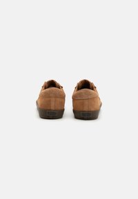 Sapatos em camurça castanhos com parte traseira arredondada, apresentando uma superfície texturizada e uma sola de borracha escura. Design minimalist sem cordões ou adornos visíveis.