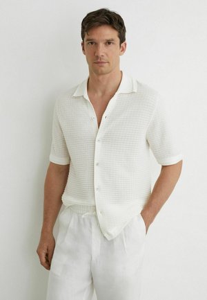 Mężczyzna o krótkich brązowych włosach nosi białą koszulę z krótkim rękawem na guziki o fakturowanej powierzchni oraz białe spodnie, stoi na tle gładkiej białej ściany.