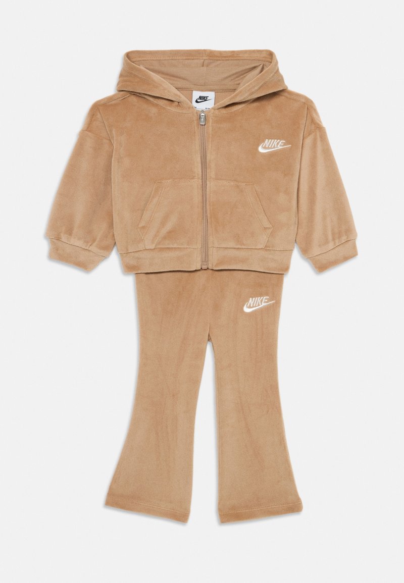 Sada z tanového velúru, ktorá obsahuje mikinu a rozšírené nohavice. Mikina má zips a predné vrecká. Obe časti majú biele logo Nike.