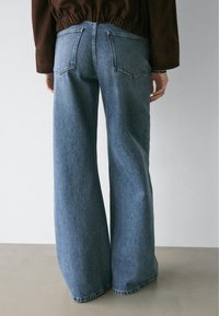 Person trägt weit geschnittene blaue Jeans und ein braunes langärmliges Oberteil, steht vor einer schlichten hellgrauen Wand.