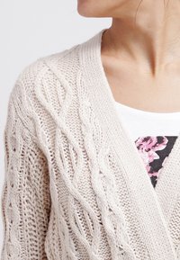 Žena v krémovém kardiganu s pleteným vzorem, přez oděv bílou košili s černým a růžovým květinovým vzorem, zachycena od ramene po krk.