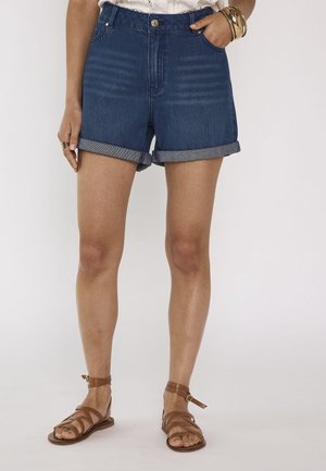 Femme portant un short en jean bleu retroussé, des sandales marron à brides, des bracelets et un haut blanc, debout devant un fond uni.