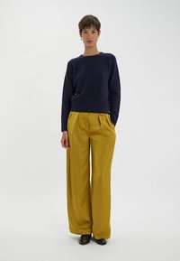 Pull en tricot bleu marine avec ourlet côtelé, associé à un pantalon large taille haute couleur moutarde. Le mannequin porte des chaussures noires. Design simple, sans motifs.