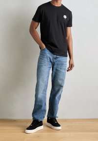Black cotton t-shirt with a small graphic on the chest, paired with light blue denim jeans and black suede sneakers featuring white soles.
