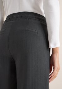 Zwarte visgraatbroek met een elastische tailleband, twee achterzakken en een gladde textuur, gecombineerd met een witte langemouwen top.