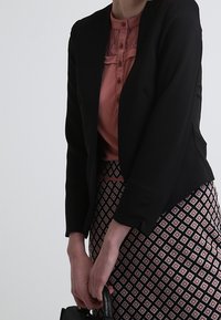 Blazer nero sopra una blusa rosa con dettagli in pizzo. Gonna a matita con motivo caratterizzato da forme geometriche audaci in rosso e bianco. Borsa nera in mano.