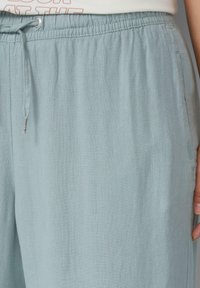 Pantalons en lin bleu clair avec une taille élastique, fermeture à cordon de serrage et poches latérales. Le tissu a une apparence douce et texturée.