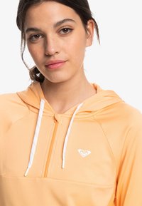 Ljust orange zip-up hoodie med vita dragningar och en liten logotyp på bröstet. Slät textur och sportig design.