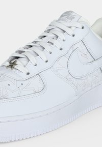 Λευκό αθλητικό παπούτσι Nike Air Force 1 με ανάγλυφα σχέδια αστεριών, λευκά κορδόνια και μικρό μεταλλικό τσόκερ κορδονιών με αστέρι.