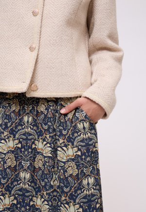 Mano in tasca, giacca beige strutturata con bottoni abbinata a una gonna blu navy decorata con intricati motivi floreali e di viti in tonalità terrose tenui.