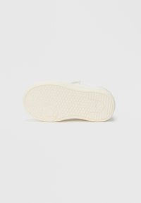 Semelle d'une sneaker blanche avec un motif en losange et un logo circulaire embossé près du talon, présentée sur un fond blanc uni.