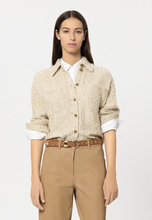 Beige textured cardigan with a collared neck, featuring gold buttons. Worn over a white shirt, paired with brown pants and a matching belt.