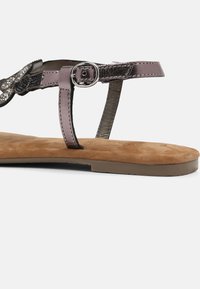 Flat sandal featuring a suede insole, metallic gray straps, and a decorative buckle. The sole is light brown with a minimal design.