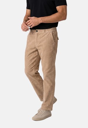 Uomo che indossa pantaloni di velluto a coste beige, sneaker beige e una maglietta nera a maniche corte, in piedi davanti a uno sfondo semplice.