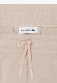 Beige elastische tailleband met trekkoord aan de broek, voorzien van een wit Lacoste-label en een klein groen krokodillenlogo.
