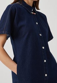 Mörkblå denimskjorta med spetsig krage, korta ärmar och knäppning fram. Har silverfärgade knappar och dekorativa pärldetaljer.