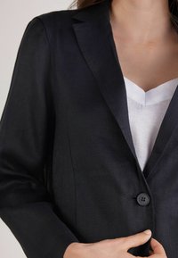 Ciemnoniebieska marynarka z lnu z klapami, z pojedynczym czarnym zapięciem na guzik i dopasowanym krojem, noszona na białej bluzce.