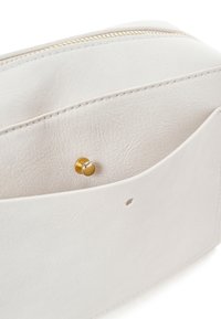 Interior de bolso de cuero blanco que muestra un bolsillo con cierre de botón metálico dorado y detalles de costura visibles.