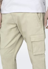 Beige cargo broek met een losse pasvorm, voorzien van meerdere zakken, een zachte textuur en subtiele stikseldetails. Zichtbare tailleband met lussen.