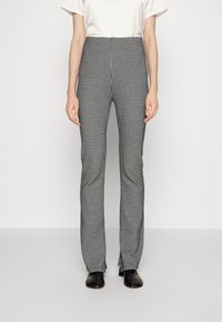Persona que lleva pantalones rectos de estampado de pied de gallo en blanco y negro, con aberturas frontales, combinados con una blusa blanca y zapatos negros.