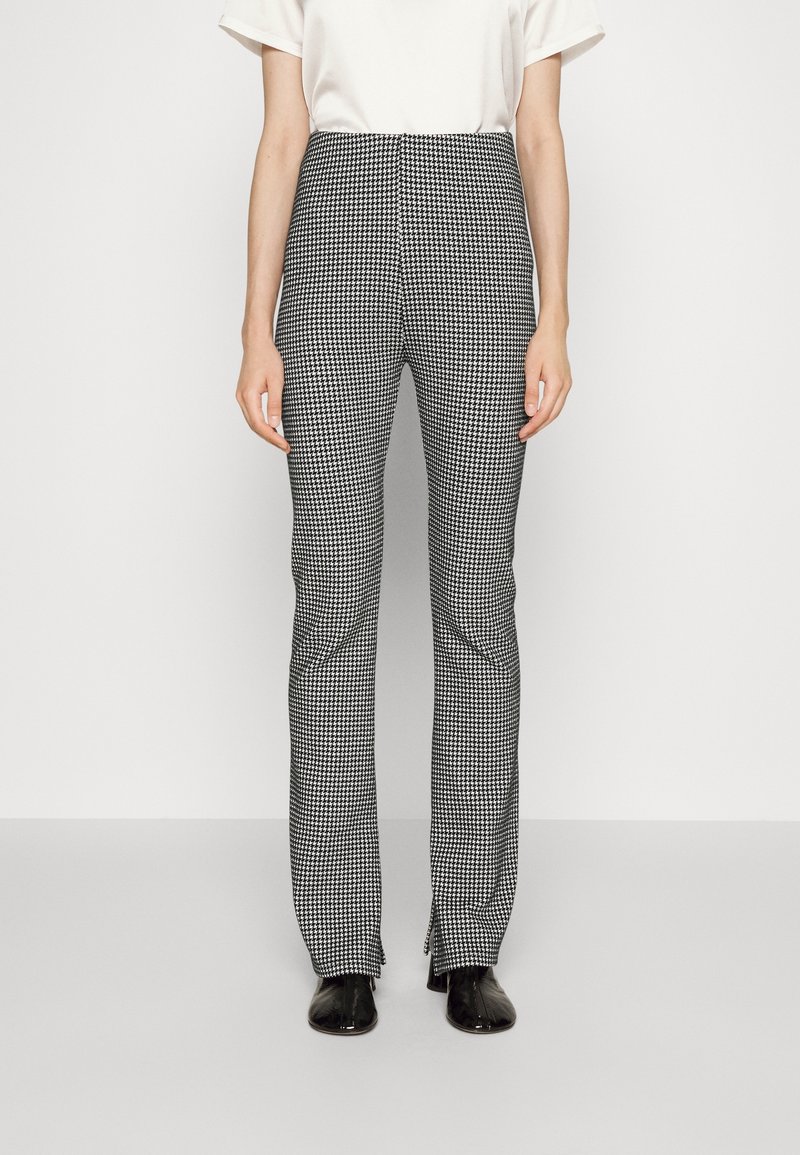 Persona que lleva pantalones rectos de estampado de pied de gallo en blanco y negro, con aberturas frontales, combinados con una blusa blanca y zapatos negros.