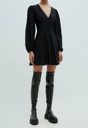 Kobieta ubrana w czarną sukienkę zapinaną na guziki z długimi bufiastymi rękawami oraz czarne masywne kozaki na platformie sięgające ponad kolano, stojąca na jednolitym tle.