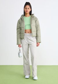 Cappotto imbottito corto verde oliva, top croppato verde chiaro, pantaloni beige con cordoncino e sneaker bianche. Piccola borsa bianca con catena.