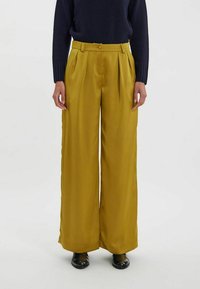Pantalons larges jaunes avec une texture douce, présentant un pli avant, une taille avec passants de ceinture et des poches latérales. Portés avec un pull bleu marine.