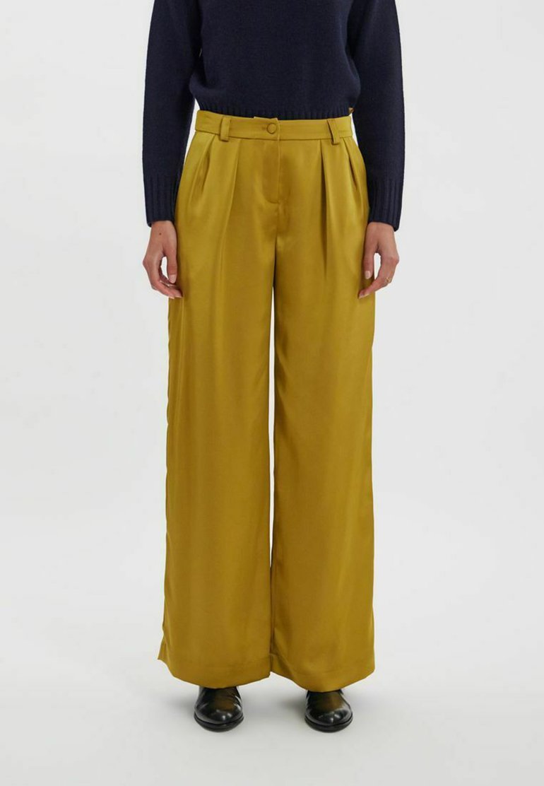 Pantalons larges jaunes avec une texture douce, présentant un pli avant, une taille avec passants de ceinture et des poches latérales. Portés avec un pull bleu marine.