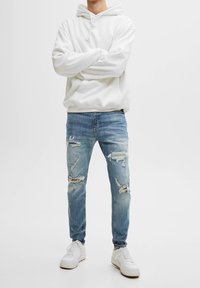 Person som bär en vit hoodie, trasiga ljusblå jeans och vita sneakers, står med armarna i kors mot en enkel bakgrund.