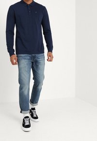 Man som bär en marinblå långärmad polo, ljusblå uppvikta jeans, vita strumpor och svarta lågskor med vita snörning.