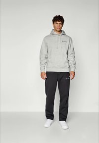 Ein junger Mann steht in einem hellgrauen Kapuzenpullover, schwarzen Jogginghosen mit Logos und weißen Sneakers vor einem schlichten hellen Hintergrund.