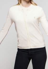 Krémově zbarvený cardigan z měkké látky, s kulatým výstřihem, zapínáním na knoflíky a malým vyšitým logem na prsou.