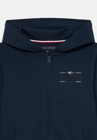 Σκούρα μπλε ζακέτα με φερμουάρ και κουκούλα, με κεντημένα τα "Hilfiger New York 1985" και με μικρό κόκκινο, λευκό και μπλε λογότυπο σημαίας στο στήθος.