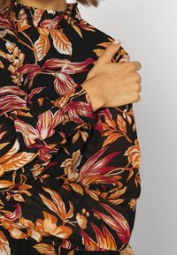 Musta kukkakuosiset paita, jossa on kirkkaat oranssit ja kermaiset lehväkuviot. Rypistynyt kaula-aukko ja kerätyt hihansuut lisäävät rakennetta ja yksityiskohtia.