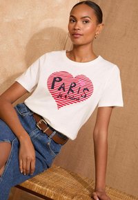 Balta medvilnės marškinėliai su raudonais dryžuotais širdies grafika, kurioje užrašyta "PARIS" ir "je t'aime" drąsiu juodu šriftu. TrumpomisRankovėmis, apvalia iškirpte.