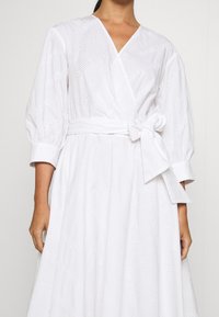 Robe blanche à manches bouffantes avec ceinture à nouer et détails en œillets. Présente un décolleté en V et une texture lisse. Agrémentée d'un motif subtil.