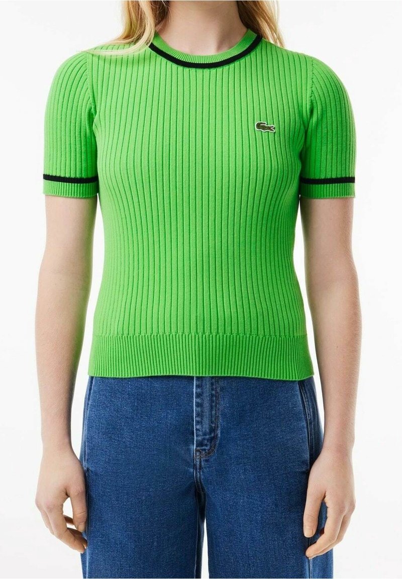 Femme portant un haut ajusté vert vif à manches courtes côtelé avec bordures noires et un petit logo crocodile, associée à un jean bleu.
