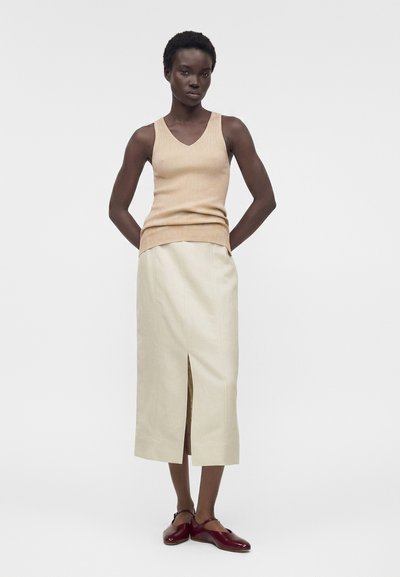 Γυναίκα που φοράει μπεζ αμάνικη ριμπ πλεκτή μπλούζα, κρεμ midi φούστα με σκίσιμο μπροστά και μπορντό παπούτσια Mary Jane, στέκεται μπροστά από απλό φόντο.