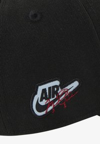 Svart caps med en rund form. Har en hvit brodert "AIR" logo og røde signaturdetaljer. Teksturert stoff med synlig søm.