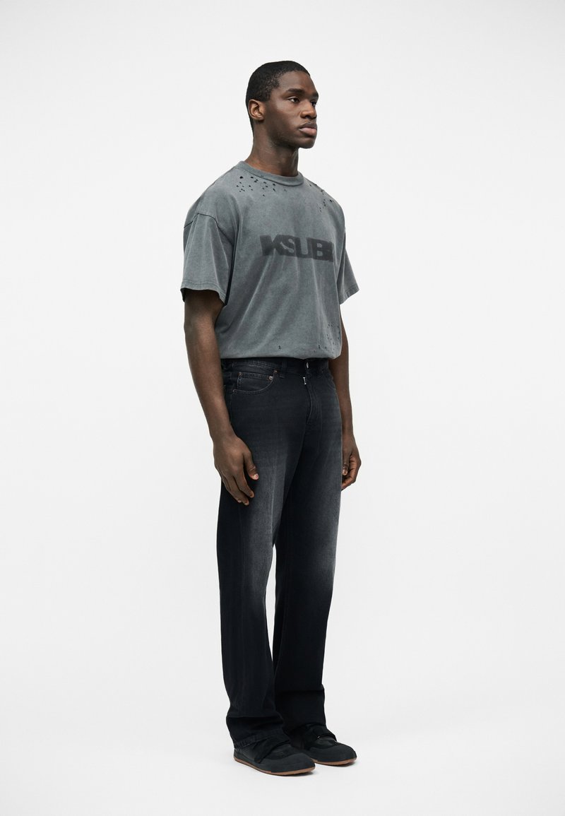 Hombre joven de pie mirando hacia adelante, vistiendo una camiseta gris descolorida de gran tamaño con texto negro y jeans negros de pierna recta junto con zapatos negros.