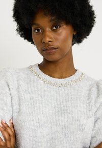 Suéter gris de punto con un acabado texturizado y mangas cortas, que presenta un cuello decorativo de cuentas con piedras transparentes.