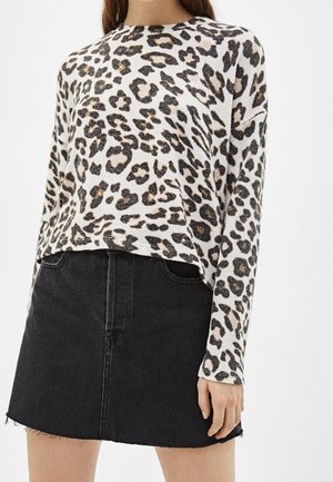 Mujer con una camiseta blanca de manga larga con estampado de leopardo y una minifalda de denim negra con dobladillo deshilachado, posando frente a un fondo liso.