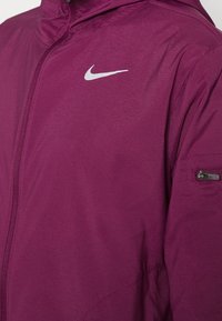 Veste de sport violette en tissu léger. Comprend une poche zippée sur la manche et un petit logo Nike blanc sur la poitrine.